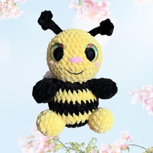 May include: A plush bee toy with a yellow body and black stripes, antennae, and wings. The bee has large, glittery eyes, a pink nose, and a smiling mouth. The toy is made of soft, textured material.