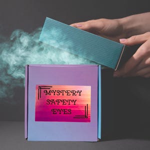 May include: A square purple box with a label that reads MYSTERY SAFETY EYES in black text. The box is partially open, with a teal lid being lifted, and a cloud of light blue smoke or vapor is emerging from the opening against a dark background.