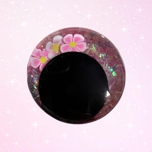 May include: A round, decorative object with a large black center. The outer ring is a mix of pink and purple with glitter and small pink flowers with yellow centers. The background is a soft pink with white sparkles.