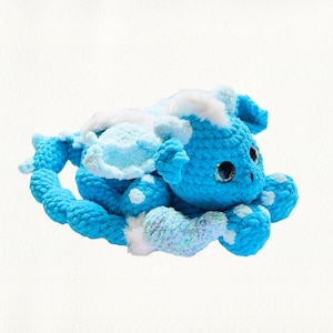 May include: A handmade blue crochet creature with white accents and large, glittery eyes. The plush toy features a long, spiraled tail and white spots on its body. The creature's wings and head are a lighter shade of blue, with white fluffy trim.