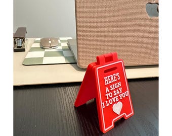May include: A red plastic sign with white text that reads "HERE'S A SIGN TO SAY I LOVE YOU" and a white heart. The sign is propped up on a black surface, with a laptop and desk accessories in the background.