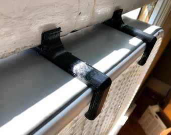 May include: Two black 3D printed hooks mounted on a white window frame. The hooks are designed to hold items like window blinds or curtains.