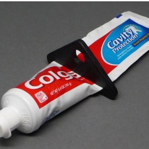 May include: A black plastic toothpaste tube squeezer with a white Colgate toothpaste tube with the text "Cavity Protection" and "Great Regular Flavor" on the tube.