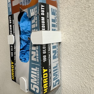 May include: A box of blue nitrile gloves with the text "Hardy 100 Gloves" and "5 mil thickness" printed on the side. The box is mounted on a white wall.