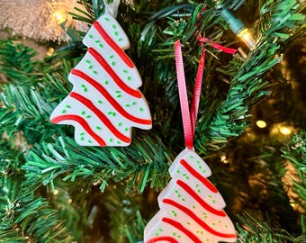 May include: Two white Christmas tree ornaments with red stripes and green dots hang from red ribbons on a green Christmas tree. The ornaments are shaped like Christmas trees.