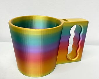 May include: A rainbow-colored mug with a unique handle design. The mug transitions through shades of gold, orange, red, purple, and teal. The handle has a wavy, ergonomic shape, and the mug appears to be 3D-printed.