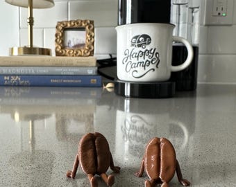 May include: Two brown coffee bean figurines with small legs are sitting on a countertop. A white mug with a camper graphic and the words "Happy Camper" sits on a coffee maker. A lamp, books, and a framed picture are in the background.