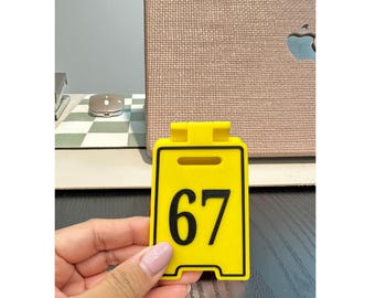 May include: A yellow sign with the number 67 in black. The sign has a rectangular shape with rounded corners and a slot at the top. The background includes a laptop and a computer mouse.