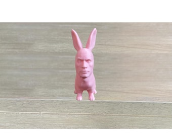 May include: A pink figurine of a rabbit with a human face. The sculpture has long ears and a detailed face. The figure is standing upright on four small feet. The color is a solid, light pink.