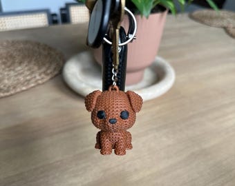 May include: A brown dog keychain with black eyes and a small nose hangs from a set of keys. The keychain is made of a textured material and is suspended in front of a wooden table. A houseplant is visible in the background.