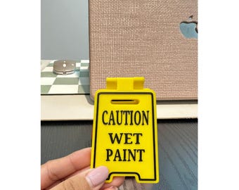 May include: A yellow and black "Caution Wet Paint" sign. The sign is rectangular with rounded corners and a handle at the top. The text is in bold, black letters. The sign is held by a person.