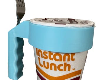May include: A light blue 3D-printed holder with a handle and a metal fork attached, designed to hold a cup of instant lunch. The cup is white with the words "Instant Lunch" in orange and a maroon stripe.