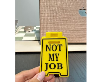 May include: A yellow sign with black text that reads "NOT MY JOB." The sign is held in a hand. The background includes a computer and a mouse on a checkered surface.
