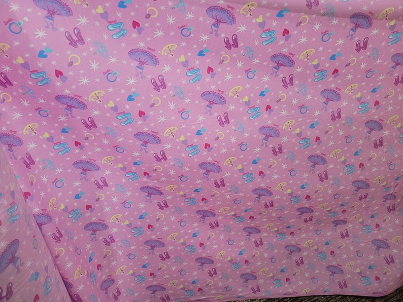 May include: Pink fabric featuring a repeating pattern of ballet shoes, umbrellas, hearts, stars, and rings. The ballet shoes are purple and blue, the umbrellas are purple and yellow, and the hearts are red and blue.