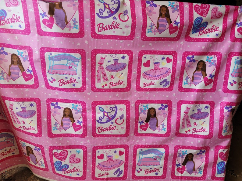May include: Pink fabric featuring repeating square patterns with Barbie illustrations. Each square contains a Barbie doll, a bed, a dress, or accessories. The word "Barbie" is written in a stylized font within each square.