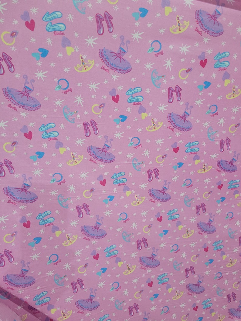 May include: Pink fabric with a repeating pattern of ballet tutus, ballet slippers, hearts, rings, and stars. The word "Barbie" is printed on some of the elements. The design features a light pink background with a variety of pastel colors.