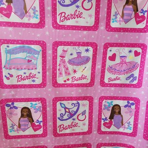 May include: Pink fabric featuring a repeating pattern of Barbie-themed squares. Each square contains illustrations of Barbie, a bed, a crown, a ring, and a tutu. The Barbie logo is present in each square. The background is a lighter pink with white star accents.