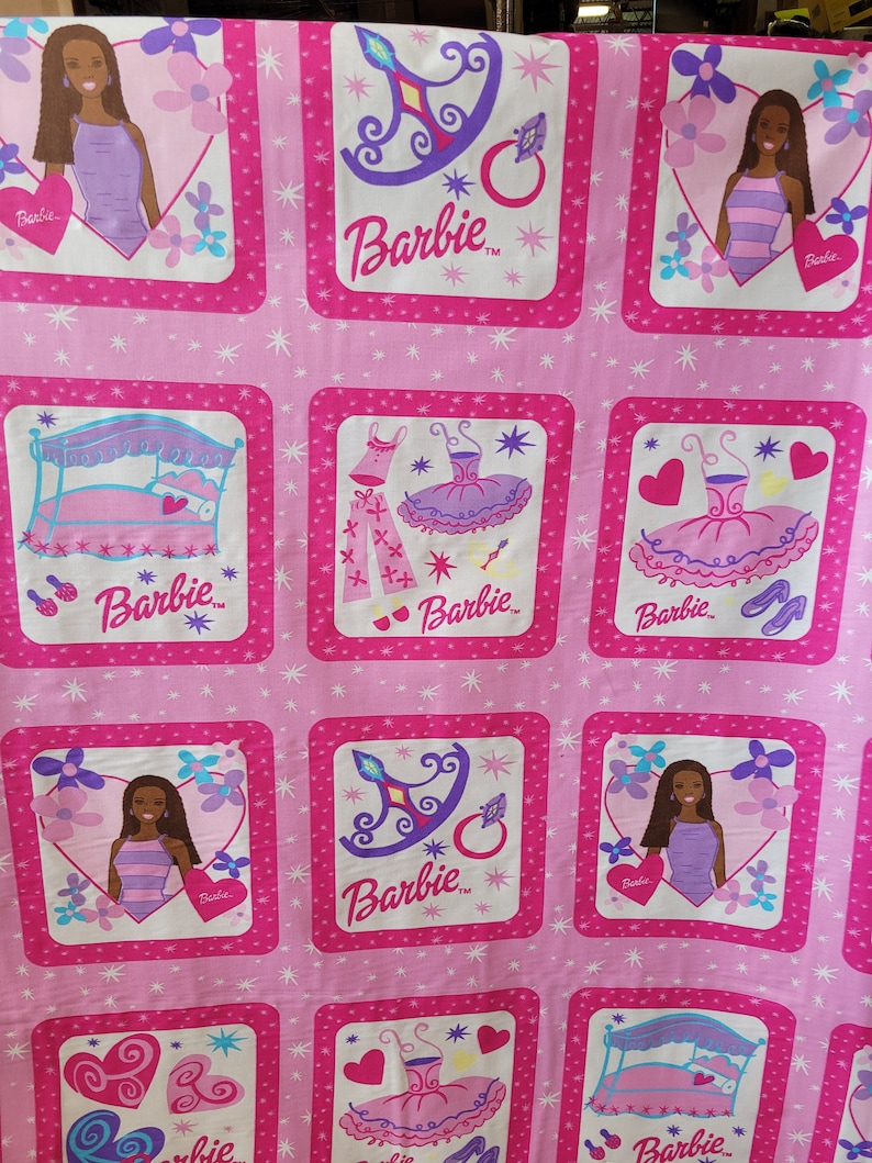 May include: Pink fabric featuring a repeating pattern of Barbie-themed squares. Each square contains illustrations of Barbie, a bed, a crown, a ring, and a tutu. The Barbie logo is present in each square. The background is a lighter pink with white star accents.