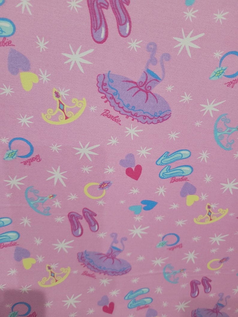 May include: Pink fabric with a repeating pattern of ballet-themed illustrations. The design includes ballet shoes, tutus, tiaras, hearts, rings, and stars. The word "Barbie" is printed on the fabric.
