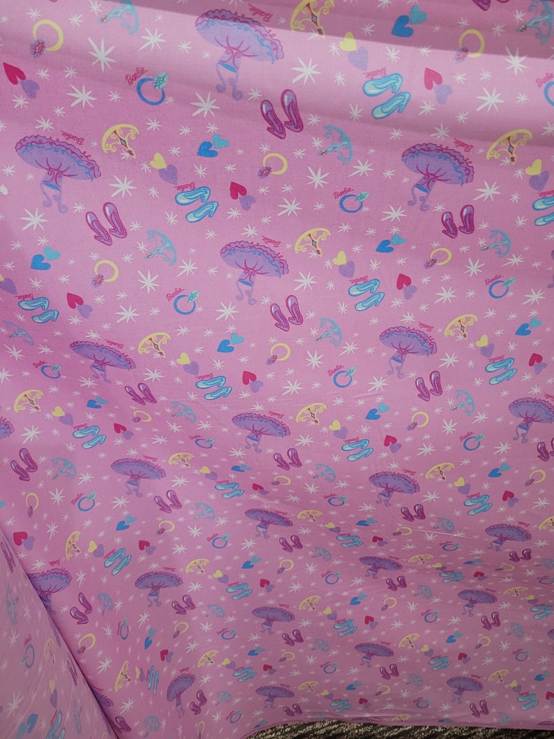 May include: Pink fabric with a repeating pattern of ballet shoes, rings, hearts, and parasols. The word "Barbie" is printed throughout the design. The fabric has a soft, flowing appearance.