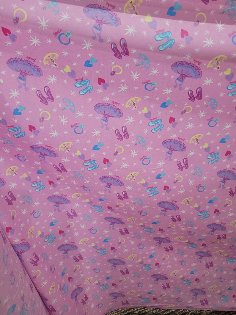 May include: Pink fabric featuring a repeating pattern of ballet shoes, parasols, hearts, rings, and stars. The design includes the word "Barbie". The overall theme is a whimsical, girly design.