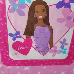 May include: Pink fabric featuring a Barbie doll illustration. The doll is inside a heart-shaped frame, surrounded by flowers. The word "Barbie" is written on a heart. The background has a pattern of stars and stripes.