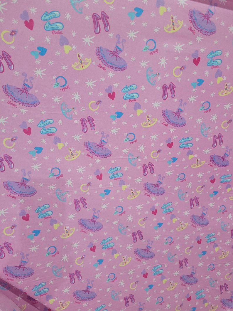 May include: Pink fabric featuring a repeating pattern of ballet dancers, ballet shoes, hearts, and stars. The word "Barbie" is printed on some of the ballet dancer images. The design includes light blue, purple, yellow, and white accents.