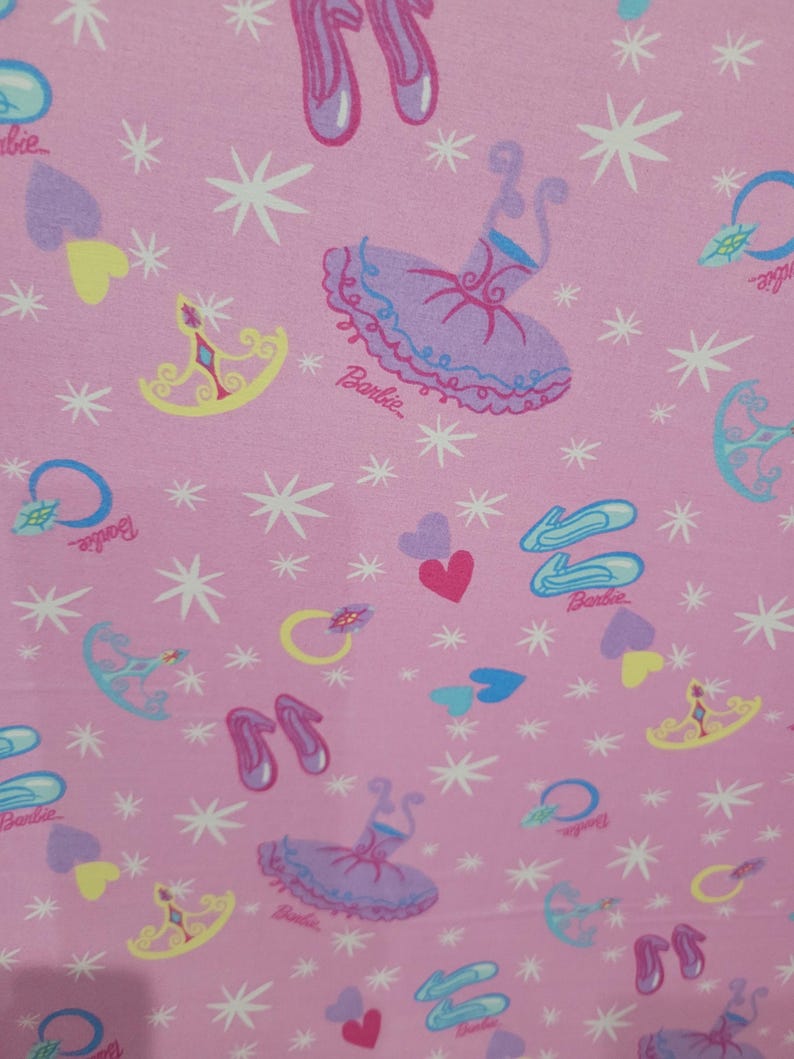 May include: Pink fabric featuring a repeating pattern of ballet shoes, tutus, hearts, crowns, rings, and stars. The word "Barbie" is printed in cursive. The design has a girly, whimsical theme.