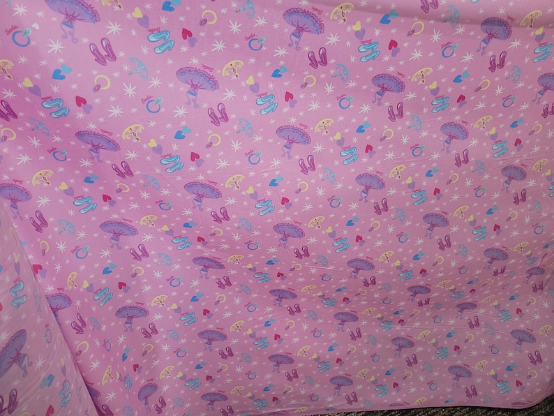 May include: Pink fabric with a repeating pattern of ballerinas, ballet shoes, hearts, rings, and stars. The ballerinas have purple tutus and are holding umbrellas. The ballet shoes are pink and white.