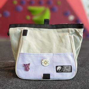 May include: A light gray climbing chalk bag with a purple pocket. The bag has a black strap and a black zipper. The pocket has a purple and black pin with a cartoon character, a silver pin with a circuit board design, and a black and white patch with the text "Further Beyond Plus Ultra".
