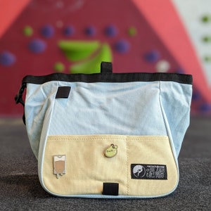 May include: A light blue and tan chalk bag for rock climbing. The bag has a black trim and a small loop at the top. The front features two pins and a patch that reads "FURTHER BEYOND + PLUS ULTRA".