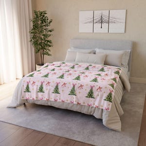 May include: A pink blanket featuring a repeating pattern of green Christmas trees, pink bows, and gold stars. The blanket is on a bed with white bedding. A painting of trees hangs on the wall.