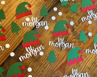 May include: Green and red elf hat cutouts with the text "hi morgan" on a wooden surface. There are also green Christmas tree cutouts and white circles.