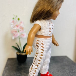 Op de afbeelding: Een witte gehaakte outfit voor een pop, bestaande uit een crop top en broek met een kruislings ontwerp. De pop draagt rode sneakers.