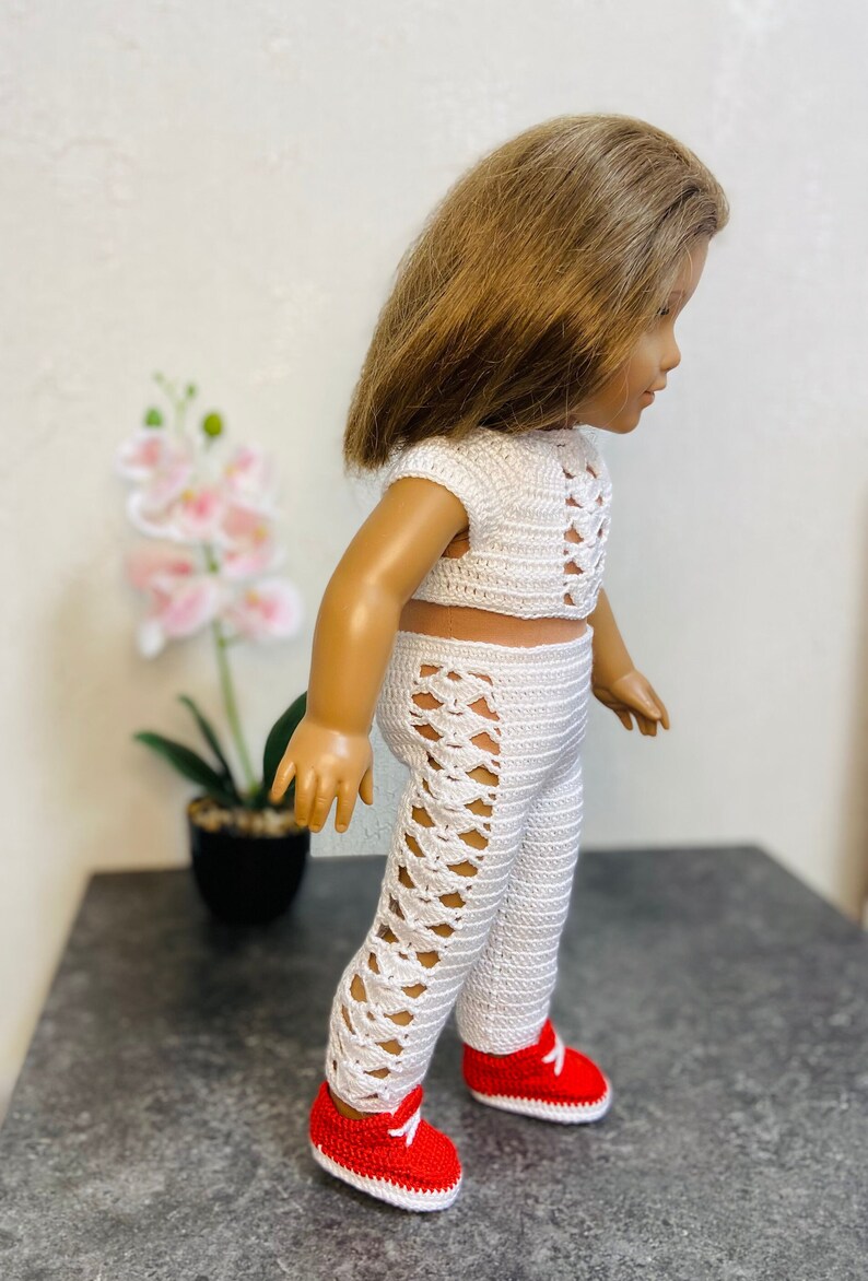 Op de afbeelding: Een witte gehaakte outfit voor een pop, bestaande uit een crop top en broek met een kruislings ontwerp. De pop draagt rode sneakers.