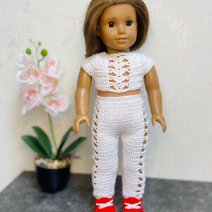 Op de afbeelding: Een wit gebreide twee-delige outfit voor een pop, bestaande uit een crop top met een veterdesign en een bijpassende broek met uitsparingen. De pop draagt rode sneakers.