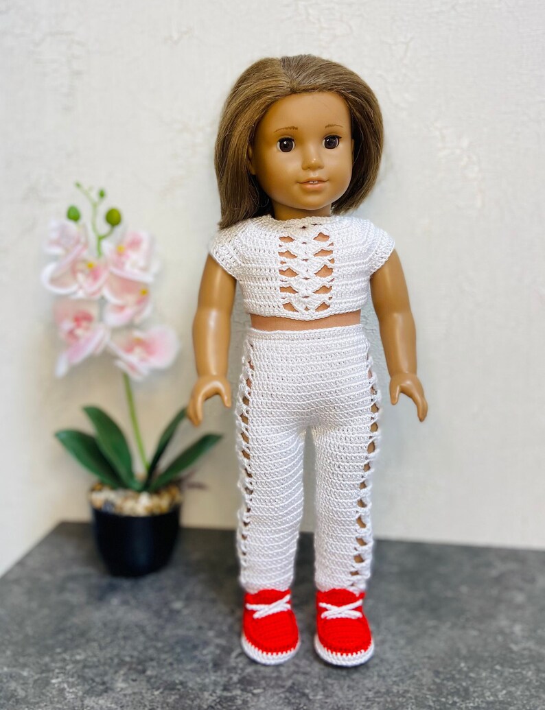 Op de afbeelding: Een wit gebreide twee-delige outfit voor een pop, bestaande uit een crop top met een veterdesign en een bijpassende broek met uitsparingen. De pop draagt rode sneakers.