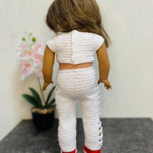 Op de afbeelding: Een wit gebreid tweedelig outfit voor een pop, bestaande uit een crop top en een broek met zijdelingse uitsnijdingen. De outfit is gemaakt met een delicaat, open steekpatroon.