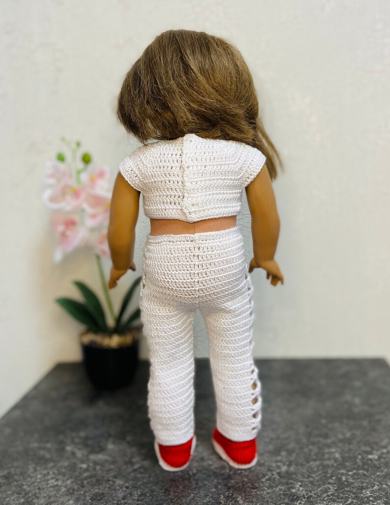 Op de afbeelding: Een wit gebreid tweedelig outfit voor een pop, bestaande uit een crop top en een broek met zijdelingse uitsnijdingen. De outfit is gemaakt met een delicaat, open steekpatroon.