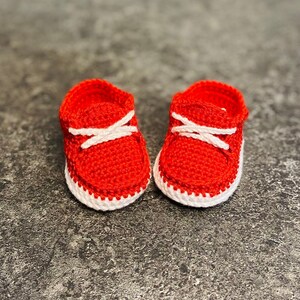 Op de afbeelding: Een paar rode gehaakte babyschoentjes met witte veters en witte rand.