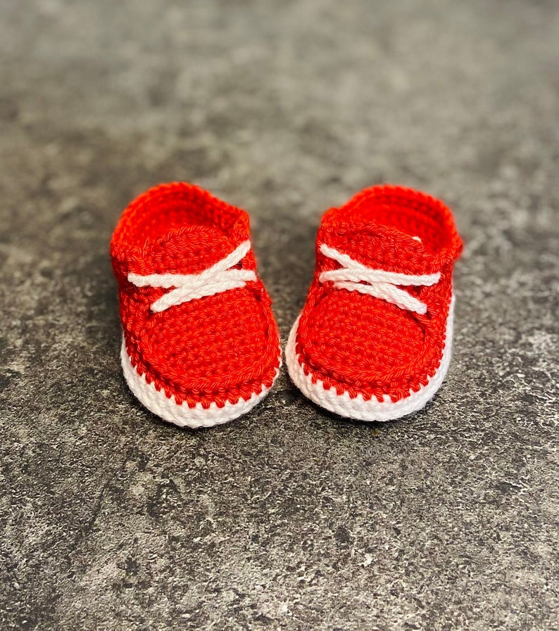Op de afbeelding: Een paar rode gehaakte babyschoentjes met witte veters en witte rand.