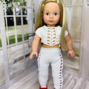 Op de afbeelding: Een witte gehaakte outfit voor een pop, bestaande uit een crop top met een veterdesign en een bijpassende broek. De pop draagt rode schoenen.