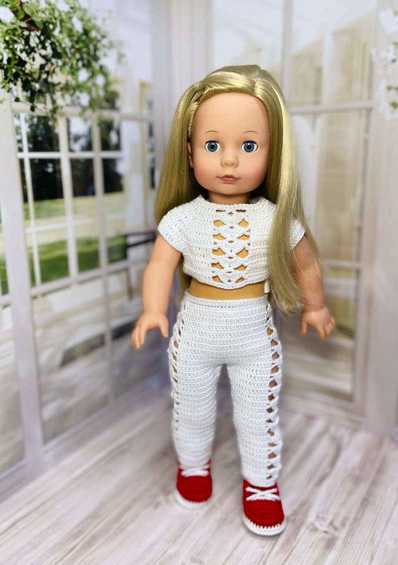 Op de afbeelding: Een witte gehaakte outfit voor een pop, bestaande uit een crop top met een veterdesign en een bijpassende broek. De pop draagt rode schoenen.