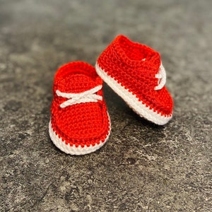Op de afbeelding: Een paar rode gehaakte babyschoentjes met witte rand. De schoentjes hebben een simpel ontwerp en zijn perfect voor een pasgeboren baby.
