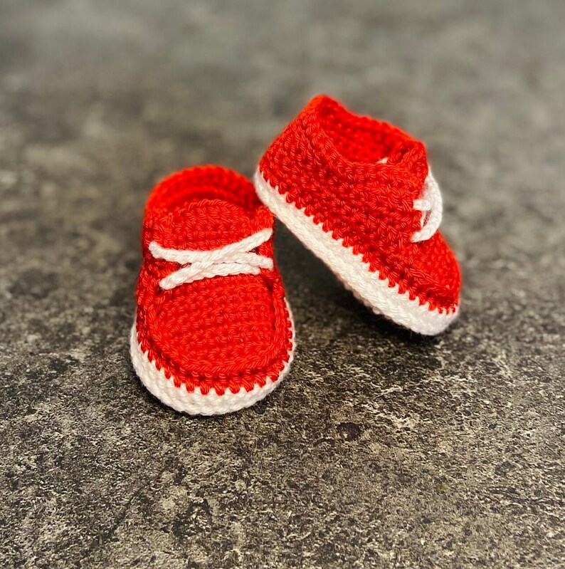 Op de afbeelding: Een paar rode gehaakte babyschoentjes met witte rand. De schoentjes hebben een simpel ontwerp en zijn perfect voor een pasgeboren baby.