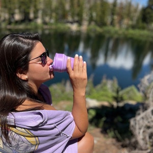 May include: A person drinking from a collapsible, lavender-colored water bottle. The bottle is being held near the person's mouth. The background features a lake and trees under a partly cloudy sky. The person is wearing sunglasses and a purple shirt.
