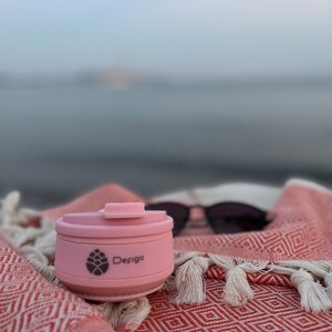May include: A pink collapsible travel mug with the logo "Defigo" and a pine cone design. The mug is sitting on a red and white patterned blanket.