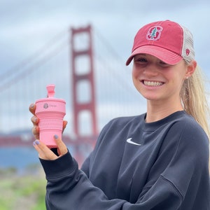 May include: A woman wearing a black sweatshirt with a white Nike swoosh logo and a red and white baseball cap with a green tree and the letter "S" on it. She is holding a pink collapsible silicone cup with the word "Stojo" printed on it. The Golden Gate Bridge is in the background.