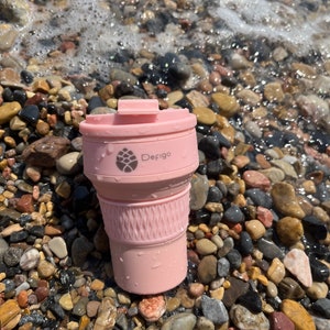 May include: A collapsible pink travel coffee cup with a lid, sitting on a bed of colorful pebbles near the water's edge. The cup has a textured grip and the brand name "Defigo" is printed on the side.
