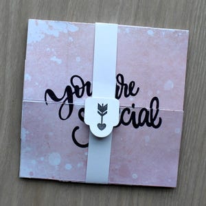 May include: A square card with a light pink watercolor-style background and the black cursive text "you are special" printed across the center. A white paper band wraps around the middle, featuring a small shield-shaped tab with a dark arrow icon.